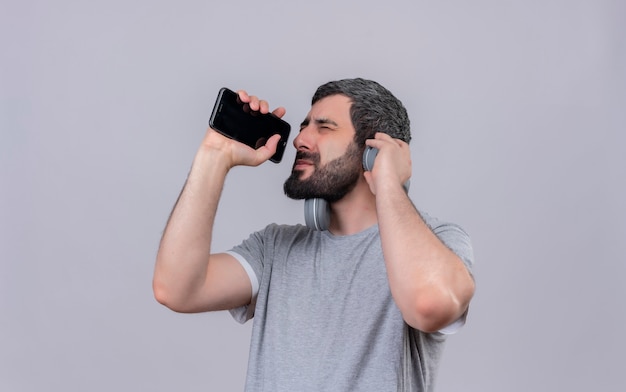 Joven apuesto hombre caucásico con auriculares finge cantar y usar su teléfono móvil como micrófono con los ojos cerrados y la mano en los auriculares aislados sobre fondo blanco con espacio de copia