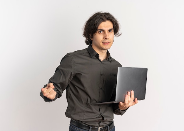 Joven apuesto hombre caucásico confiado sostiene portátil y apunta a la cámara aislada sobre fondo blanco con espacio de copia