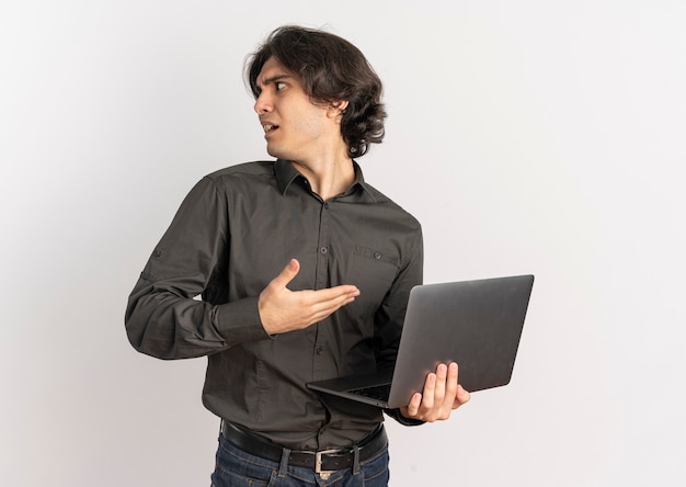 Joven apuesto hombre caucásico molesto sostiene y apunta a la computadora portátil mirando al lado aislado sobre fondo blanco con espacio de copia