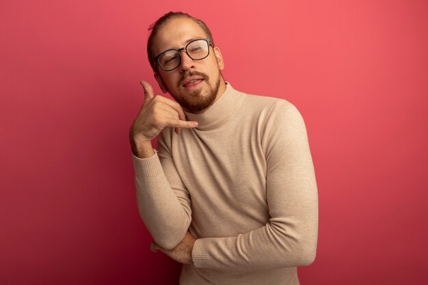 Joven apuesto hombre de cuello alto beige y gafas mirando a la cámara sonriendo confiado haciendo gesto de llamarme de pie sobre la pared rosa