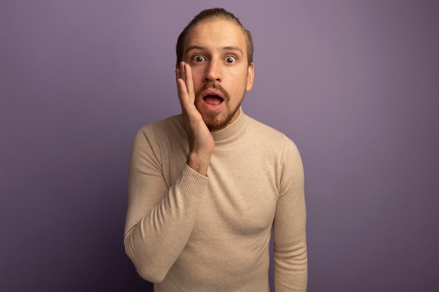 Joven apuesto hombre de cuello alto beige mirando al frente con la mano ner boca sorprendida de pie sobre la pared lila