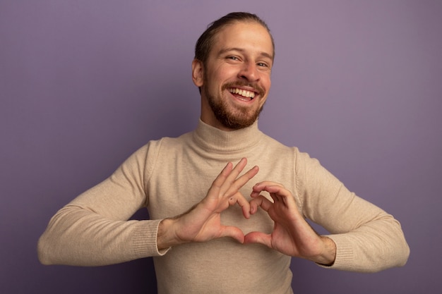 Foto gratuita joven apuesto hombre de cuello alto beige mirando al frente sonriendo alegremente haciendo gesto de corazón con los dedos de pie sobre la pared lila