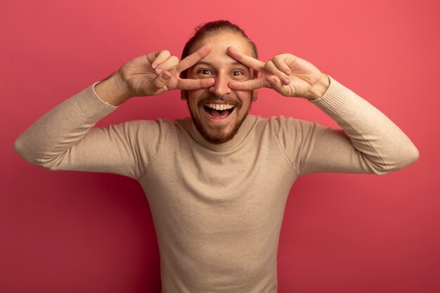 Joven apuesto hombre de cuello alto beige mirando al frente sonriendo alegremente mostrando v-sign de pie sobre la pared rosa