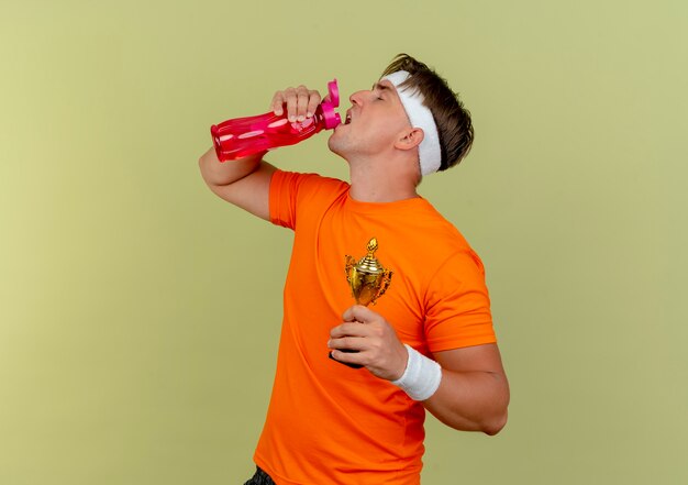 Joven apuesto hombre deportivo con diadema y muñequeras sosteniendo la taza del ganador y bebiendo agua de la botella de agua aislada sobre fondo verde oliva con espacio de copia