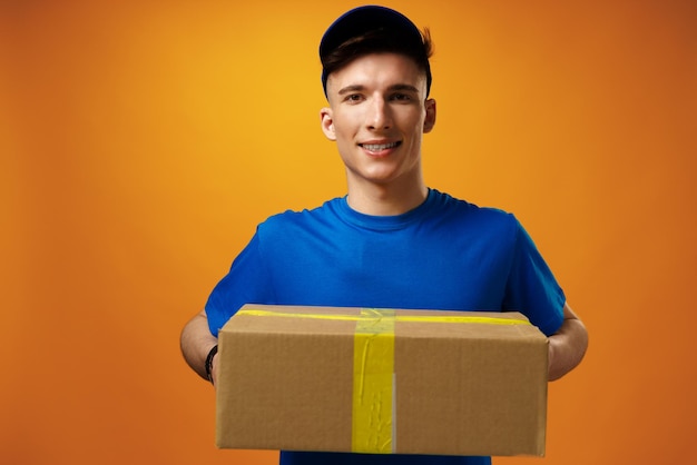 Foto gratuita joven apuesto hombre de entrega que sostiene la caja de cartón del paquete contra el fondo amarillo