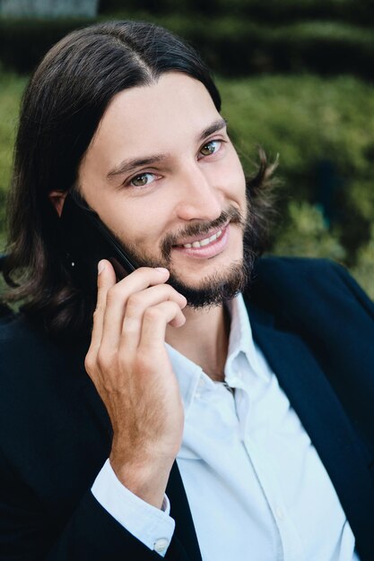 Joven apuesto hombre de negocios barbudo confiado mirando alegremente a la cámara mientras habla por teléfono celular en el restaurante al aire libre