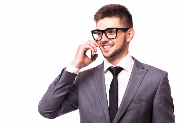 Joven apuesto hombre de negocios con camisa blanca y anteojos, hablando por teléfono y sonriendo en reacción a la broma de un colega, aislado sobre fondo gris