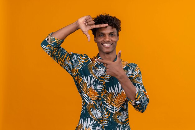 Un joven apuesto hombre de piel oscura con cabello rizado en hojas de camisa estampada haciendo marco con manos y dedos con cara feliz