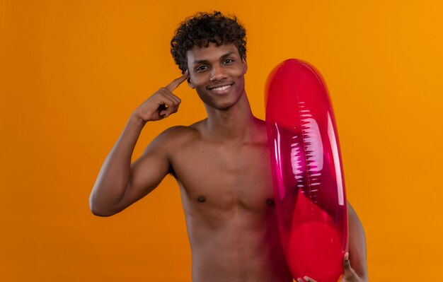 Un joven apuesto hombre de piel oscura positivo con cabello rizado apuntando con el dedo índice mientras sostiene el anillo de piscina inflable