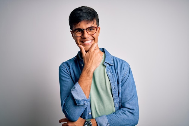 Foto gratuita joven apuesto hombre vestido con camisa informal y gafas sobre fondo blanco aislado mirando confiado a la cámara sonriendo con los brazos cruzados y la mano levantada en la barbilla pensando en positivo