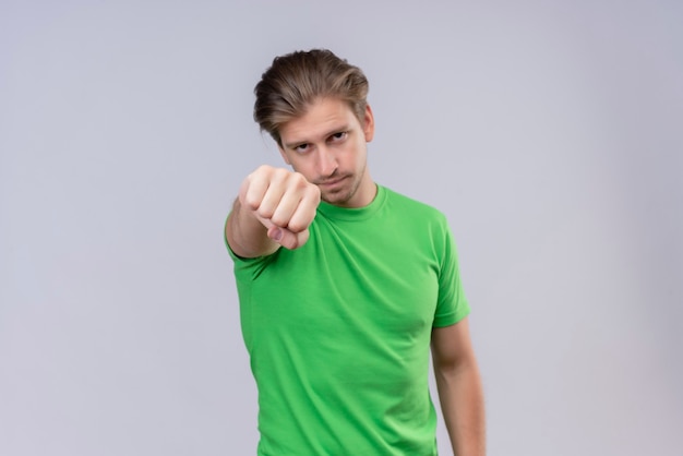 Joven apuesto hombre vestido con camiseta verde apretando el puño que se extiende a la cámara con la cara seria de pie sobre la pared blanca