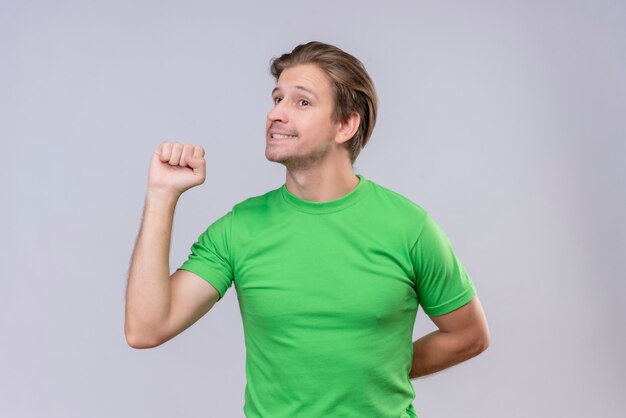 Joven apuesto hombre vestido con camiseta verde apretando el puño sonriendo feliz y positivo de pie sobre la pared blanca