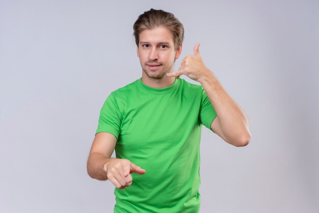 Joven apuesto hombre vestido con camiseta verde haciendo gesto de llamarme sonriendo amable de pie sobre la pared blanca