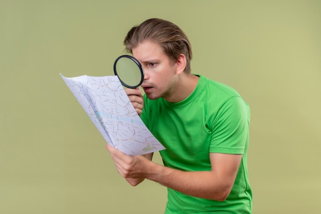 Foto gratuita joven apuesto hombre vestido con camiseta verde mirando el mapa a través de una lupa sorprendido de pie sobre la pared verde