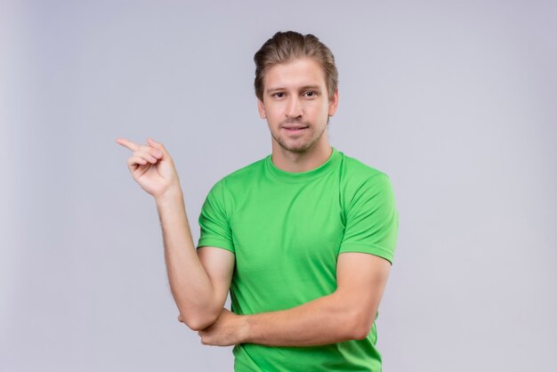 Joven apuesto hombre vestido con camiseta verde sonriendo y apuntando con el dedo índice hacia el lado