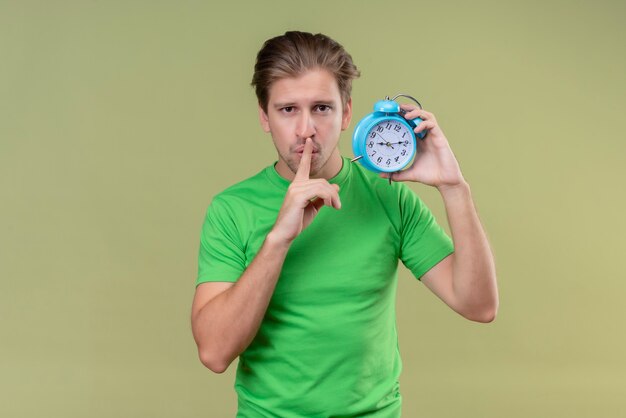 Joven apuesto hombre vestido con camiseta verde sosteniendo despertador haciendo gesto de silencio con el dedo en los labios de pie sobre la pared verde