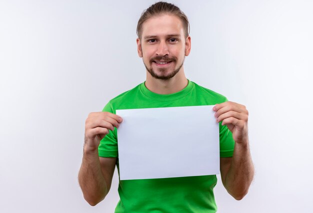 Joven apuesto hombre vestido con camiseta verde sosteniendo papel en blanco mirando a la cámara sonriendo confiado de pie sobre fondo blanco.