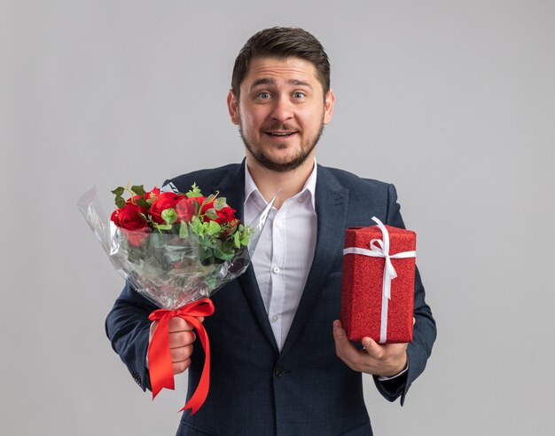 Joven apuesto hombre vestido con traje sosteniendo un ramo de rosas y un regalo para el día de San Valentín con cara feliz sonriendo alegremente de pie sobre la pared blanca