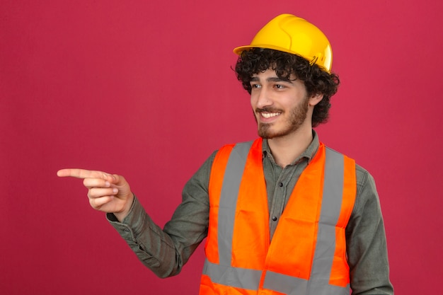 Joven apuesto ingeniero barbudo con casco de seguridad y chaleco mirando a un lado apuntando a algo con el dedo sonriendo sobre pared rosa aislado