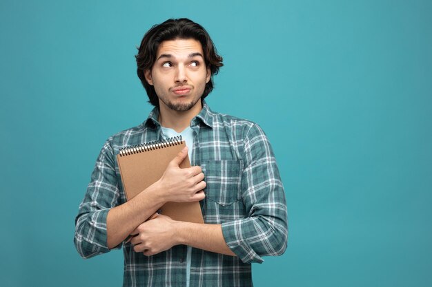 un joven apuesto inseguro abrazando un bloc de notas mirando a un lado aislado en un fondo azul con espacio para copiar