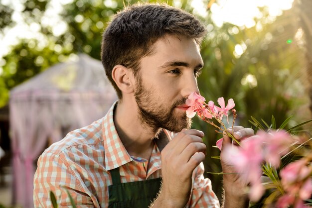 Joven apuesto jardinero alegre sonriendo, oliendo flores rosas