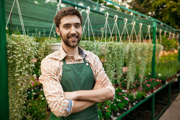 Foto gratuita joven apuesto jardinero alegre sonriendo, posando con los brazos cruzados entre flores