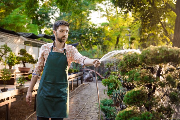 Joven apuesto jardinero alegre sonriendo, regando, cuidando las plantas