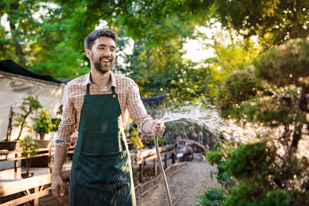 Joven apuesto jardinero alegre sonriendo, regando, cuidando las plantas