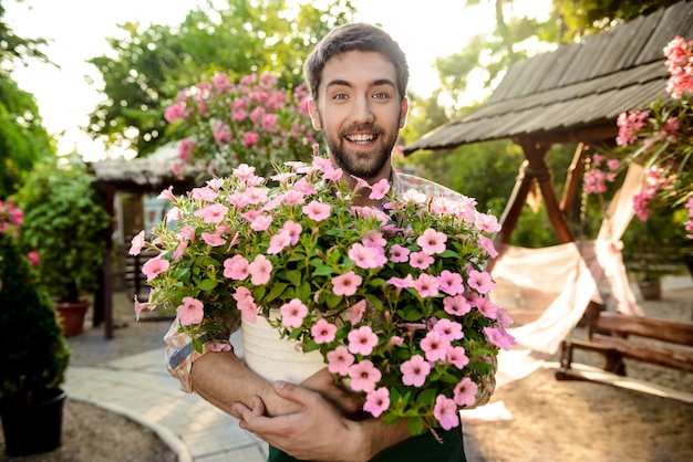 Joven apuesto jardinero alegre sonriendo, sosteniendo una olla grande con flores