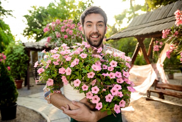 Joven apuesto jardinero alegre sonriendo, sosteniendo una olla grande con flores