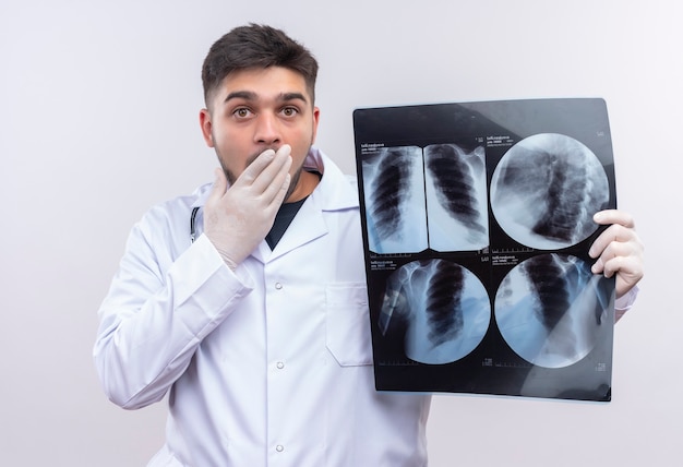 Joven apuesto médico con bata médica blanca, guantes médicos blancos y estetoscopio asustado sosteniendo tomografía de pie sobre una pared blanca