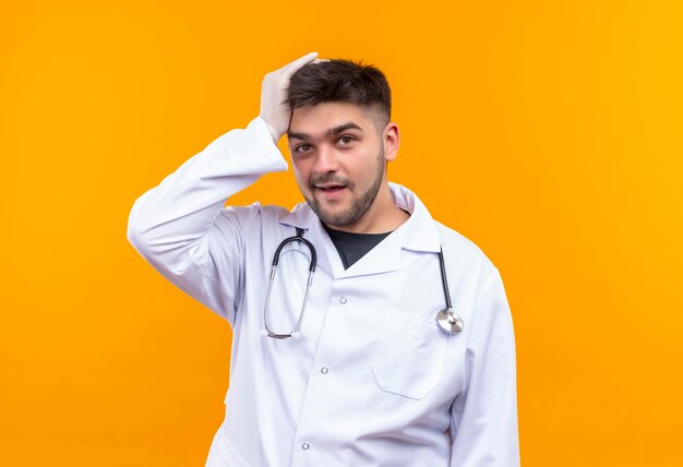 Joven apuesto médico con bata médica blanca, guantes médicos blancos y estetoscopio en la cabeza con la mano mirando con la cara olvidada de pie sobre la pared naranja