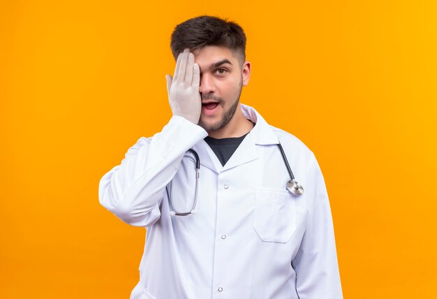 Joven apuesto médico con bata médica blanca, guantes médicos blancos y un estetoscopio cerrando un ojo de pie sobre la pared naranja