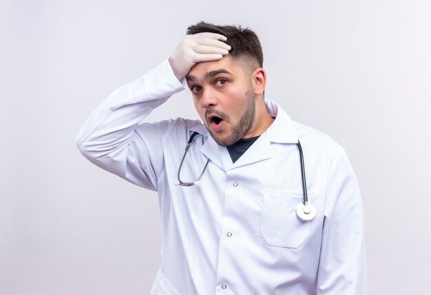 Joven apuesto médico con bata médica blanca, guantes médicos blancos y estetoscopio conmocionado de pie sobre la pared blanca