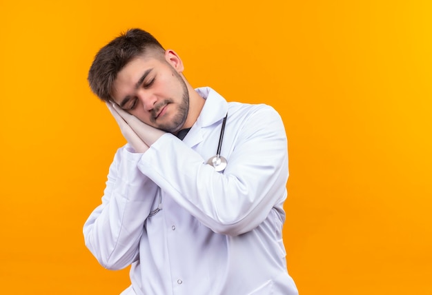 Joven apuesto médico con bata médica blanca, guantes médicos blancos y estetoscopio durmiendo de pie sobre la pared naranja