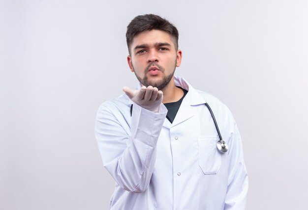 Joven apuesto médico con bata médica blanca, guantes médicos blancos y estetoscopio enviando besos de pie sobre la pared blanca