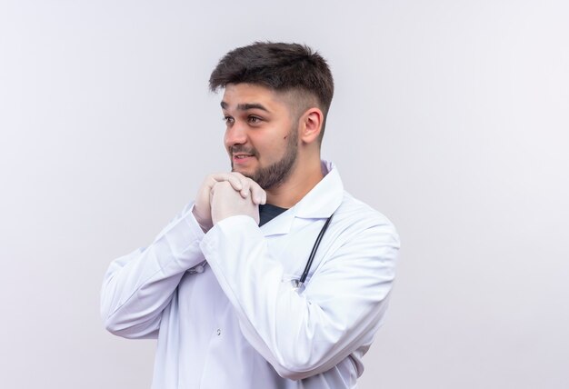 Joven apuesto médico con bata médica blanca, guantes médicos blancos y estetoscopio mirando además de fascinado parado sobre la pared blanca