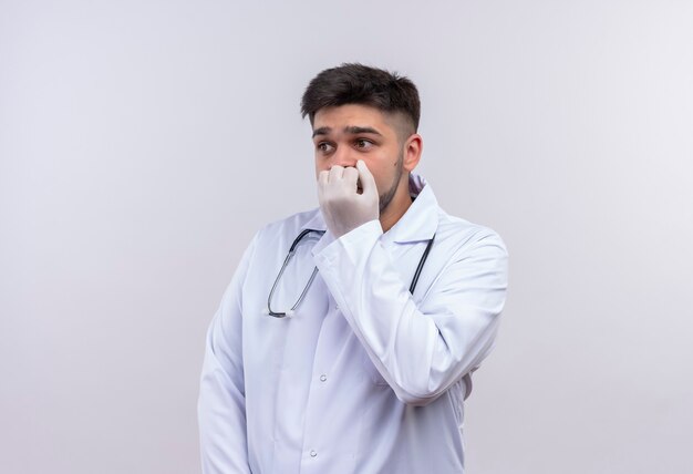 Joven apuesto médico con bata médica blanca, guantes médicos blancos y un estetoscopio mirando asustado además de sostener el puño en el ratón de pie sobre la pared blanca