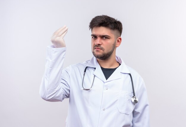 Joven apuesto médico con bata médica blanca, guantes médicos blancos y un estetoscopio mirando con enojo de pie sobre la pared blanca