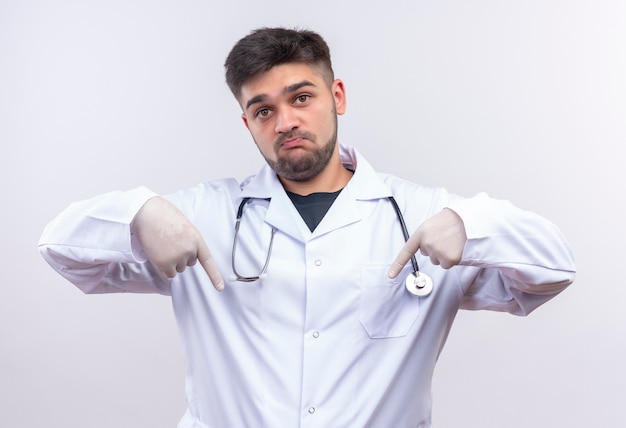 Foto gratuita joven apuesto médico con bata médica blanca guantes médicos blancos y un estetoscopio mirando molesto apuntando hacia abajo con los dedos índices de pie sobre la pared blanca