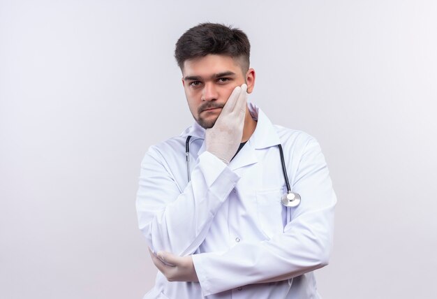 Joven apuesto médico con bata médica blanca, guantes médicos blancos y un estetoscopio mirando pensativamente de pie sobre la pared blanca