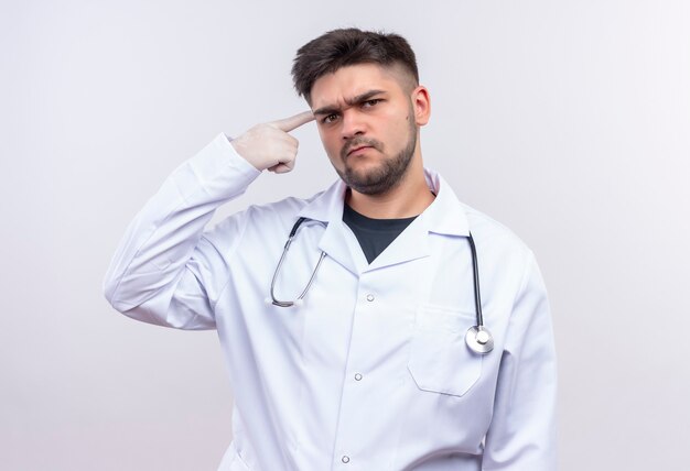 Joven apuesto médico con bata médica blanca, guantes médicos blancos y estetoscopio mirando con reproche haciendo quiere que alguien piense de pie sobre una pared blanca