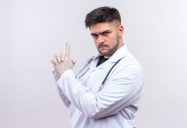 Joven apuesto médico con bata médica blanca, guantes médicos blancos y estetoscopio mirando seriamente haciendo pistola con las manos de pie sobre la pared blanca