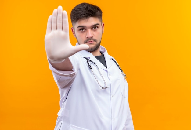 Joven apuesto médico con bata médica blanca, guantes médicos blancos y un estetoscopio mirando seriamente mostrando la señal de stop con la mano derecha de pie sobre la pared naranja