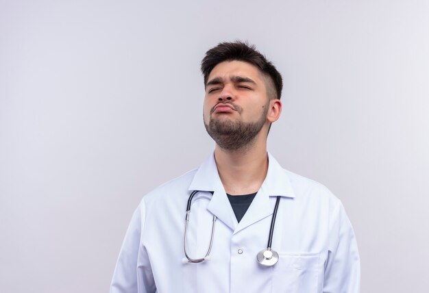 Joven apuesto médico con bata médica blanca, guantes médicos blancos y estetoscopio molesto de pie sobre la pared blanca