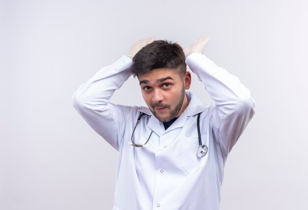 Joven apuesto médico con bata médica blanca guantes médicos blancos y un estetoscopio mostrando cuernos de pie sobre la pared blanca