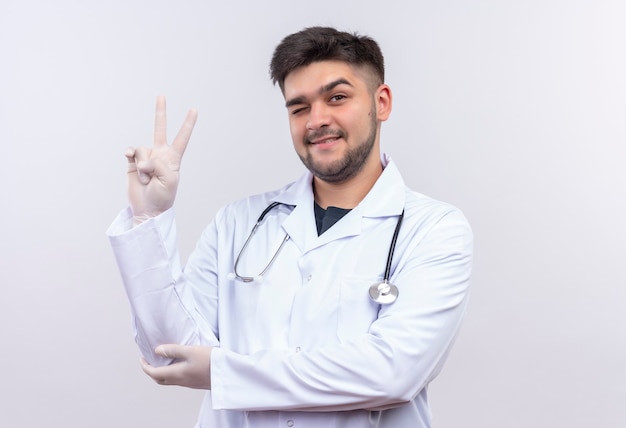 Joven apuesto médico con bata médica blanca, guantes médicos blancos y estetoscopio parpadeando un ojo mostrando el signo de la paz con los dedos de pie sobre la pared blanca