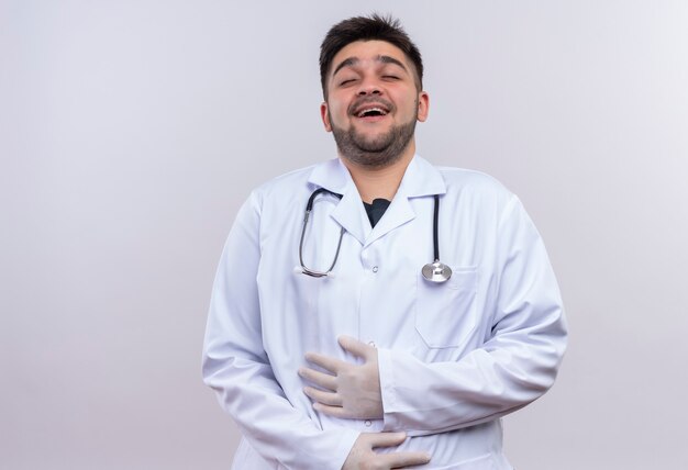 Joven apuesto médico con bata médica blanca guantes médicos blancos y un estetoscopio de pie riendo sobre la pared blanca