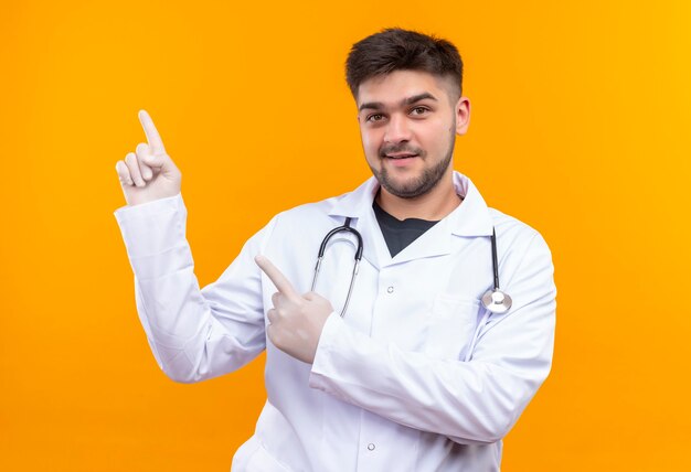 Joven apuesto médico con bata médica blanca, guantes médicos blancos y estetoscopio de pie sonriendo apuntando hacia arriba con los dedos índices sobre la pared naranja