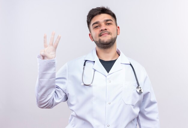 Joven apuesto médico con bata médica blanca, guantes médicos blancos y un estetoscopio que muestra con mucho gusto tres signos con los dedos de pie sobre la pared blanca